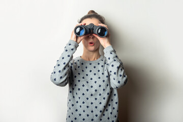 Young woman with a surprised face in a sweater looks through binoculars on a light background