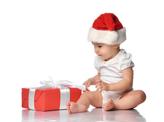 Cute infant baby girl in jumpsuit and red Santa Claus hat sitting on white floor studio background with wrapped holiday gift box with bow. Isolated portrait shot. Christmas, New Year concept