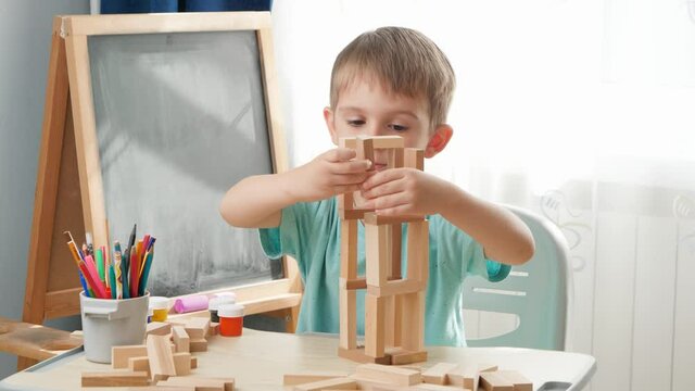 Happy smiling boy looking at toy wooden blocks tower he built on desk at school classrom. Concept of smart children and home education during lockdown and self isolation.