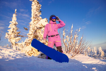 Young woman in protective helmet and ski suit holds snowboard on background of mountains sunset