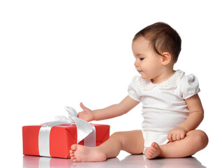 Baby and wrapped festive gift box with bow. Cute interested curious infant girl child in jumpsuit sitting on white floor studio background looking at present. Birthday or Christmas celebration concept