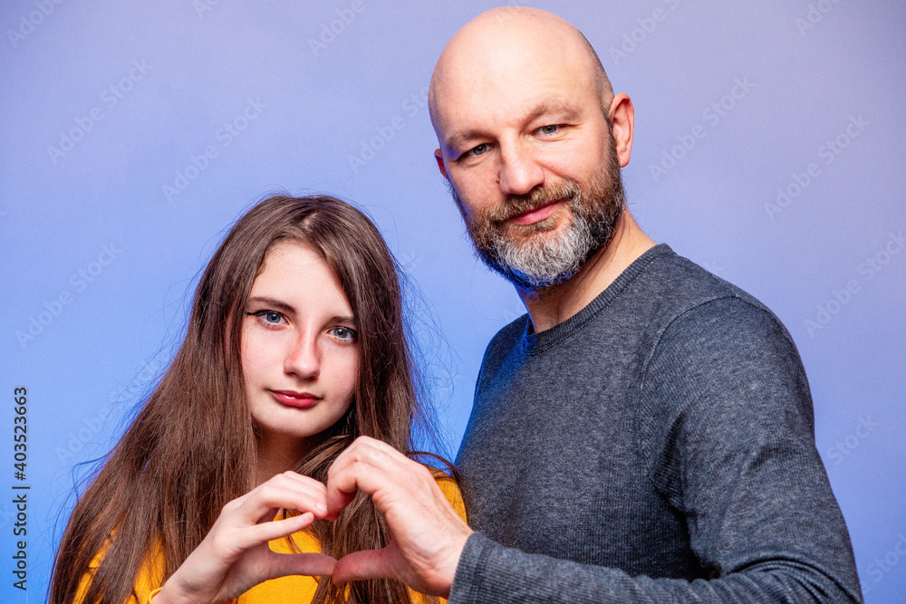 Wall mural Father and his daughter interactions. Models man in his 40s, bald with grey beard. girl teenager with long hair. Making heart looking at the camera. Blue background