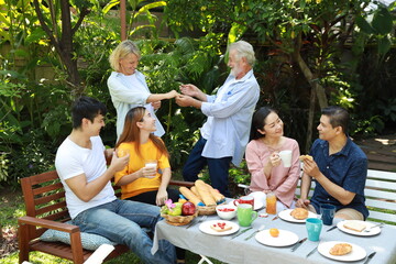Happy multiethnic family standing and sitting at a breakfast table in backyard outdoor on sunny day with smiling face. Happy caucasian and asian family sitting and having conversation with good time