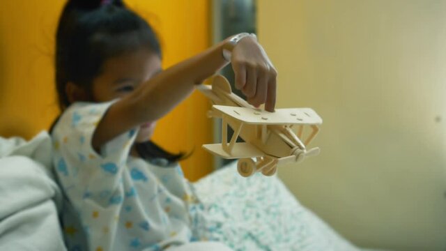 Sick asian little girl playing toy wooden airplane while stay in the hospital bed alone. Therapy for patients to relax. Recovering little girl. Slow motion shot.
