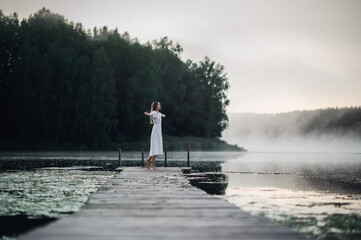 Happy woman in a white dress enjoys morning on a lake. Fog floats over the water.