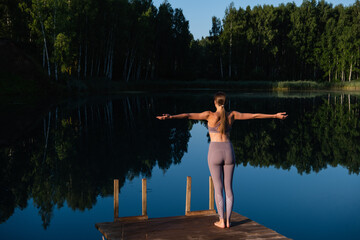 Fototapeta na wymiar Young woman walks on wooden pier above forest lake scenery, preparing for morning yoga workout in nature.