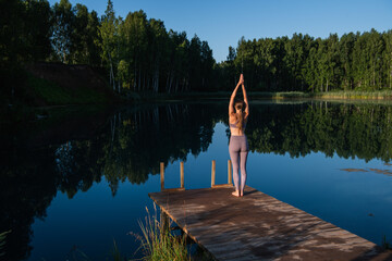 Fototapeta na wymiar Young Woman practicing yoga on the wooden berth at lake. Single sport healthy training on nature at sunny weather