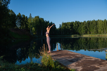 Fototapeta na wymiar Young woman relaxing by the lake at sunset, harms outstretched. Doing youga in the morning at forest lake.
