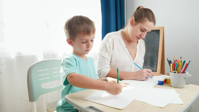 Smiling mother with little son drawing picture with pencil. Child sitting behind school desk and doing homework with parent. Education and remote school at home during lockdown and self isolation.
