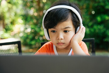 Serious looking asian kid wear headphone using laptop at yard.