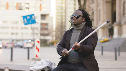 Blind black man sitting on the bench and holding walking long cane while waiting for the bus. High...