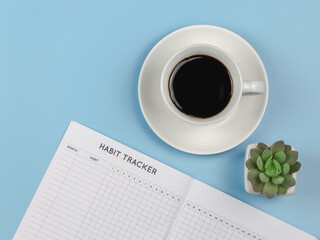 flat lay of habit tracker book with pen,  cup of black coffee and succulent plant pot on blue background with copy space.