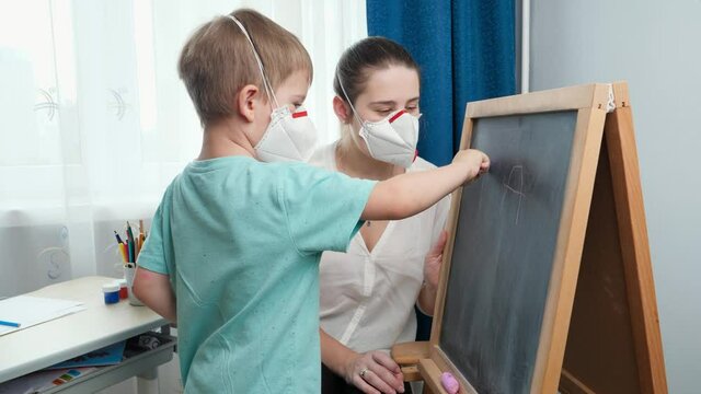 Little boy wearing protective medical mask writing letters on blackboard with chalk. Educating and learning in classrom on chalkboard during self isolation and lockdown. Remote school at home