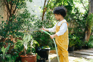 Little gardener kid watering plant with pipehose in garden.