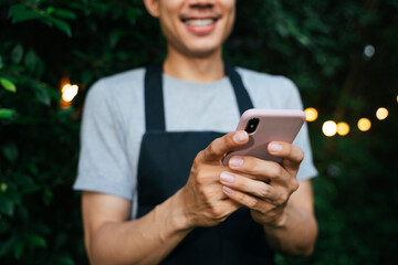 Cheerful smile asian waiter wear black apron usign smartphone at yard restaurant.