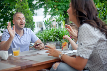 Happy people communicate in cafe. Family, mother father and daughter laughing and drink tje juice and cocktails on summer veranda of cafe