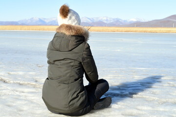 Young woman in warm clothes sitting on the ice and looking at snowy mountains. winter, Russia.