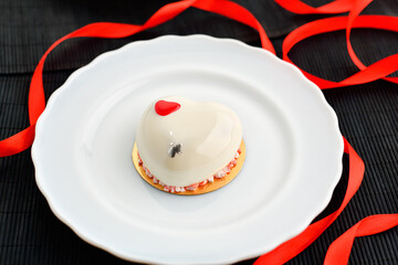 A cake in heart shape. One heart-shaped mousse cupcake on a plate on a black background with a red ribbon. Valentine's Day.