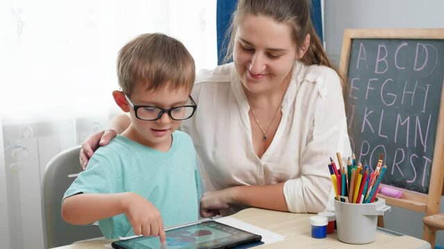 Little boy in eyeglasses playing on tablet computer. Mother hugging her little son doing homework using gadet. Children having problems with eyes and sight
