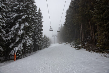 Empty Ski Lift on a snowy winter landscape, with snow covered mountains and big pine and fir trees, in the fog. The chairlift has no people on it.