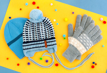 Hat and gloves on a yellow background. Clothes for the cold seasons in the form of a hat and gloves. A blue knitted hat and gray warm gloves create coziness in winter and autumn.