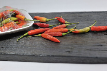 Some red chilies on a black wooden base and a bunch of red chilies in a plastic box on a white...
