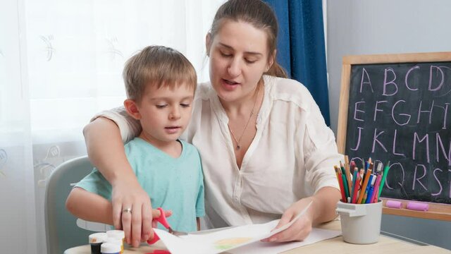 Young female teacher showing her little student how to use scissors. Boy cutting paper with scissors while doing homework.