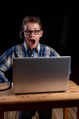 Boy with shirt and glasses joyfully with open mouth and headset doing e-learning on laptop
