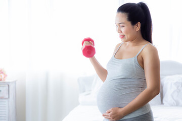 Smiling pregnant female do exercise in bedroom. Lifting dumbbells. Healthy pregnancy, sport, fitness, people and healthy lifestyle concept.