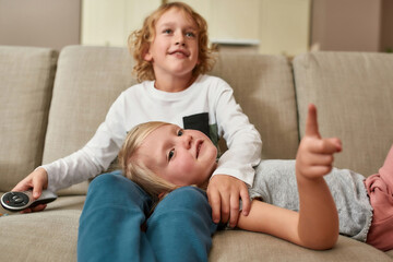 Obraz premium Portrait of cute little siblings, boy and girl looking focused while watching TV together, cuddling on a sofa at home