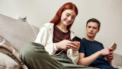 Couple of teenagers sitting on the couch at home, using mobile phone. A guy jealously looks at girlfriend phone screen. No trust, smartphone addiction, communication problem concept