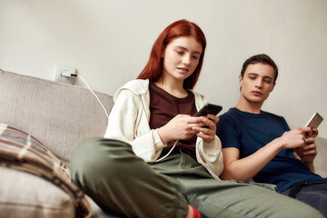 Couple of teenagers sitting on the couch at home, using mobile phone. A guy jealously looks at girlfriend phone screen. No trust, smartphone addiction, communication problem concept