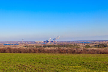 Fototapeta premium View of green wheat field and smoke pipes of factory at spring