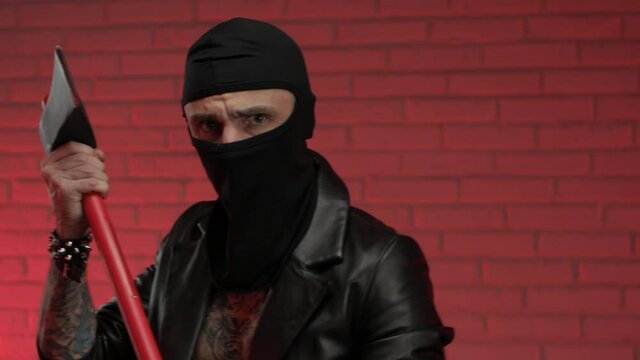 male criminal in tattoos leather raincoat in a balaclava with an axe