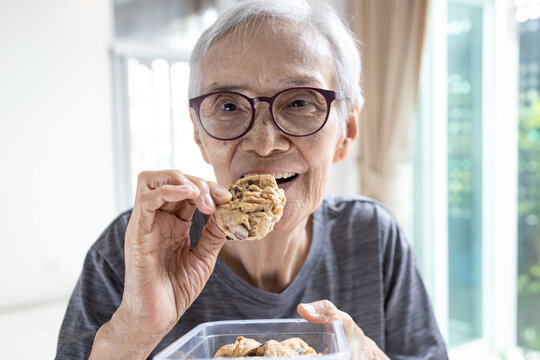 Happy Asian Senior Woman Eating Chocolate Chip Cookie Or Tasting Sweet Cookie,natural Tasty Food,delicious Snack,old Elderly Holding A Box Of Cashew Nut Biscuits In Her Hand,breakfast In The Morning