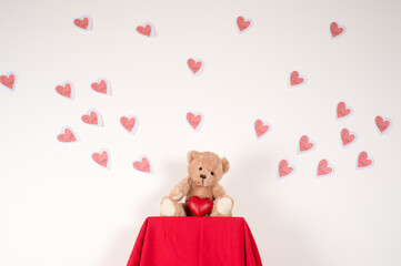 One teddy bears sitting on red fabric with one red heart in front of him, and many paper hearts flying above him on the white wall behind him