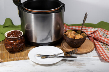 On the table is a slow cooker, a bowl with a spoon and cooked meat, a ceramic pot, and a plate with a knife and fork.
