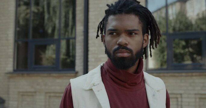 Close up view of bearded young serious guy looking aside while standing at city street. Portrait of handsome afroamerican man with dreadlocks in turtleneck posing outdoors