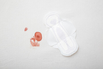 White sanitary towel and red fresh blood stain on light gray bed sheet after night menstruation. Closeup. Top down view.