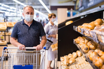 Confident buyer in protective mask with shopping cart in hypermarket