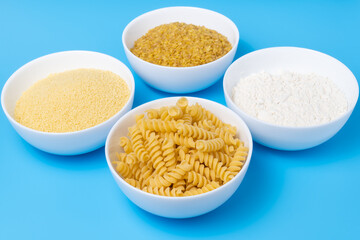 Flour, koukous, bulgur and pasta in bowls on cheerful blue background. Variety of wheat products concept.