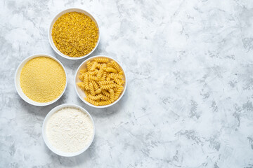 Flour, koukous, bulgur and pasta in bowls on grey textured background with space for text, top view. Various wheat products concept.