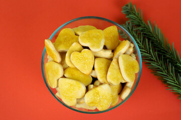 a lot of heart-shaped curd cookies smeared with orange-yellow jam on top lie in a round glass bowl next to a green Christmas tree branch on a red background on the side. homemade cakes