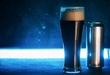 A glass of dark craft beer and an aluminum beer can. Trending neon blue backlit background, copy space