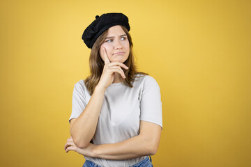 Beautiful young caucasian girl wearing french look with beret over isolated yellow background serious face thinking about question with hand on chin, thoughtful about confusing idea