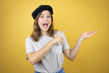 Beautiful young caucasian girl wearing french look with beret over isolated yellow background surprised, showing and pointing something that is on her hand