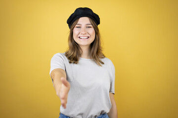 Beautiful young caucasian girl wearing french look with beret over isolated yellow background smiling friendly offering handshake as greeting and welcoming