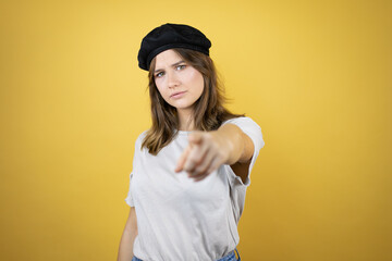 Beautiful young caucasian girl wearing french look with beret over isolated yellow background pointing with finger to the camera and to you, confident gesture looking serious