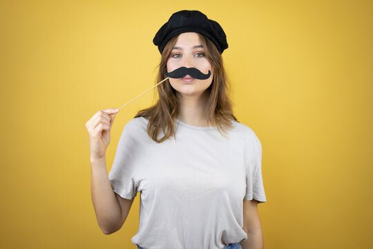 Beautiful young caucasian girl wearing french look with beret over isolated yellow background happy with mustache mask
