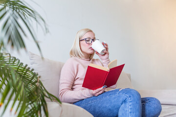 Young woman at home sitting on modern sofa relaxing in her living room reading book and drinking coffee or tea. White cozy bed and a beautiful girl, reading a book, concepts of home and comfort